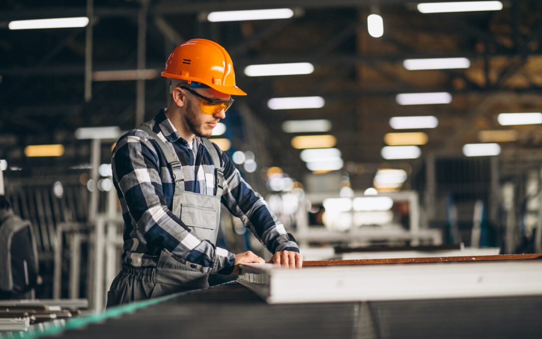 Factory worker in an orange hard hat and safety glasses checking a panel on a production line in a manufacturing hall
