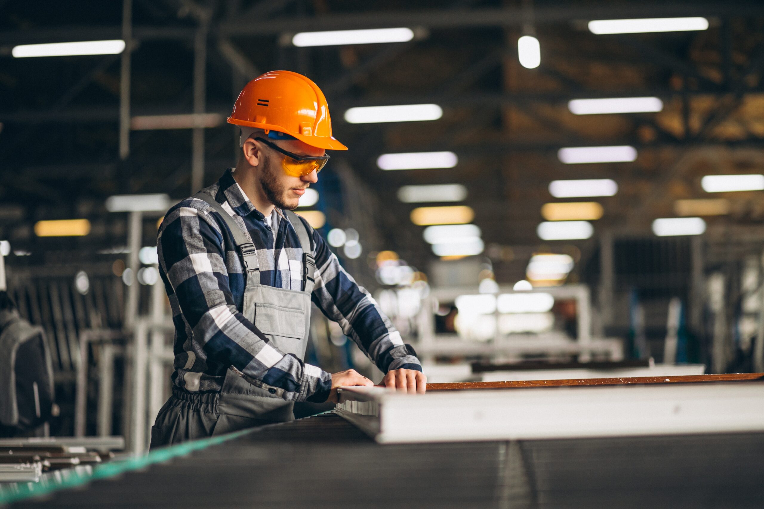 Factory worker in an orange hard hat and safety glasses checking a panel on a production line in a manufacturing hall